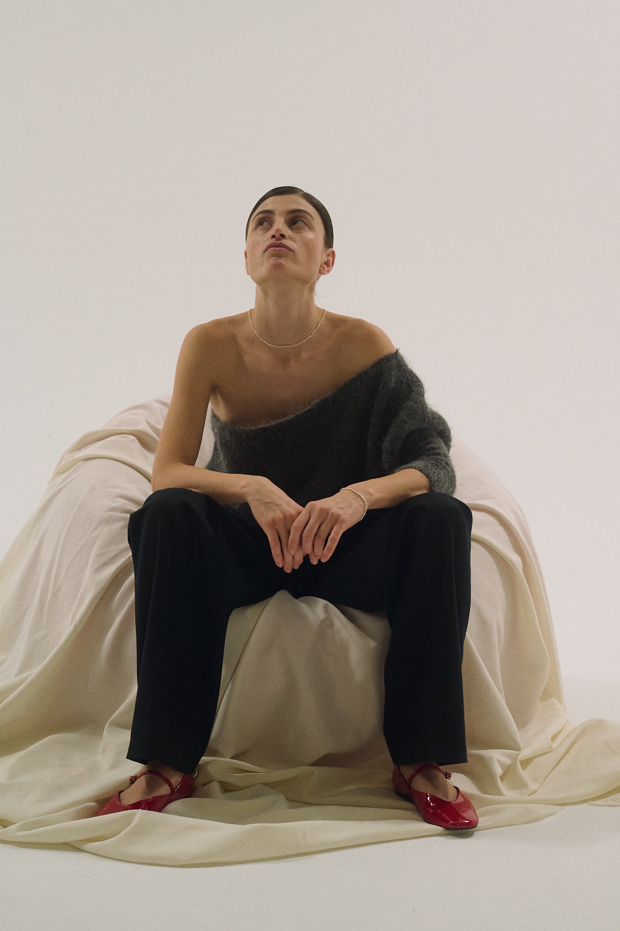 Model styling tiny pearl bracelet with relaxed outfit, sitting on fabric chair