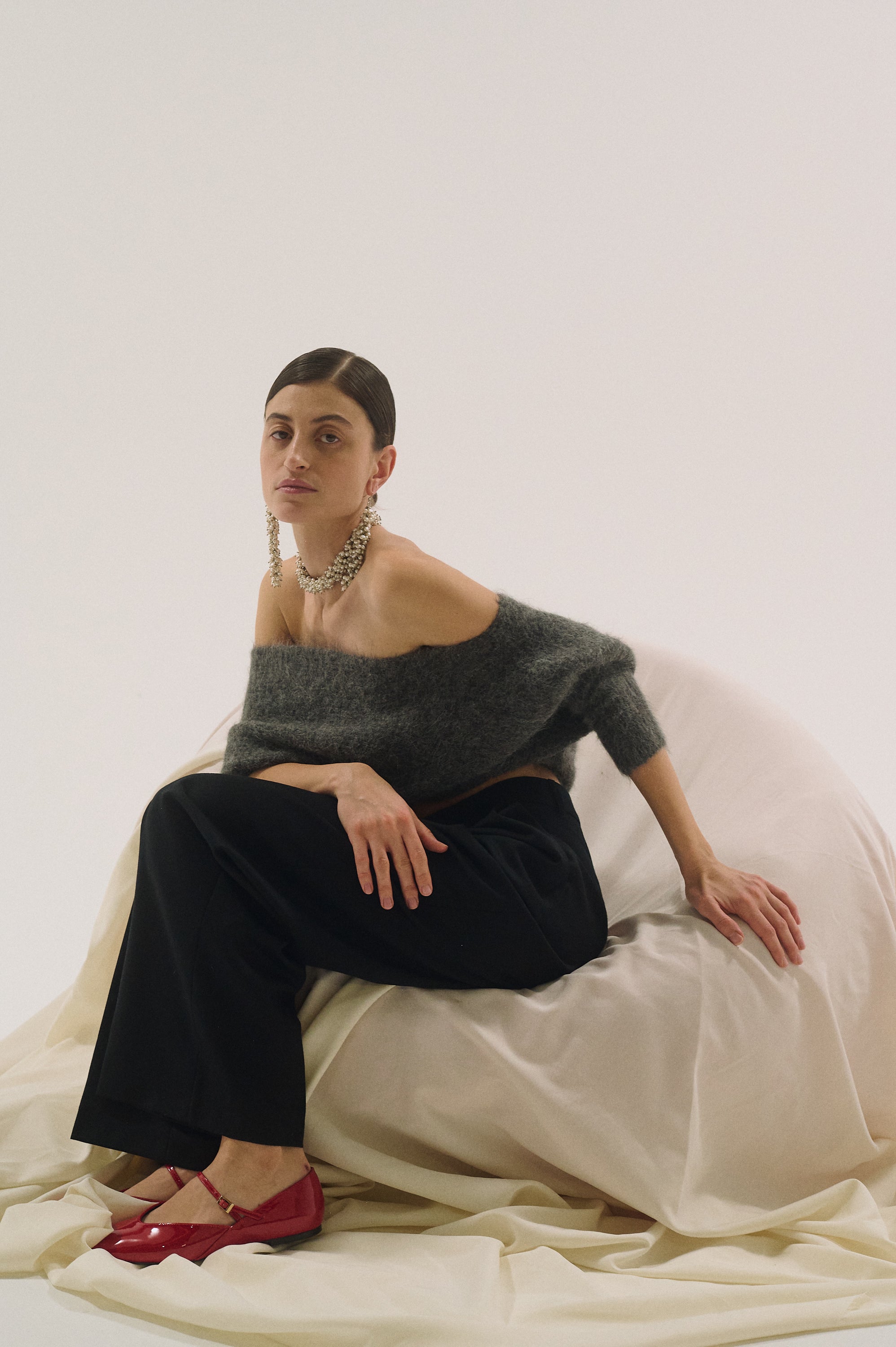 Woman sitting on a white draped fabric wearing a frop freshwater pearl earrings, gray sweater and black pants.