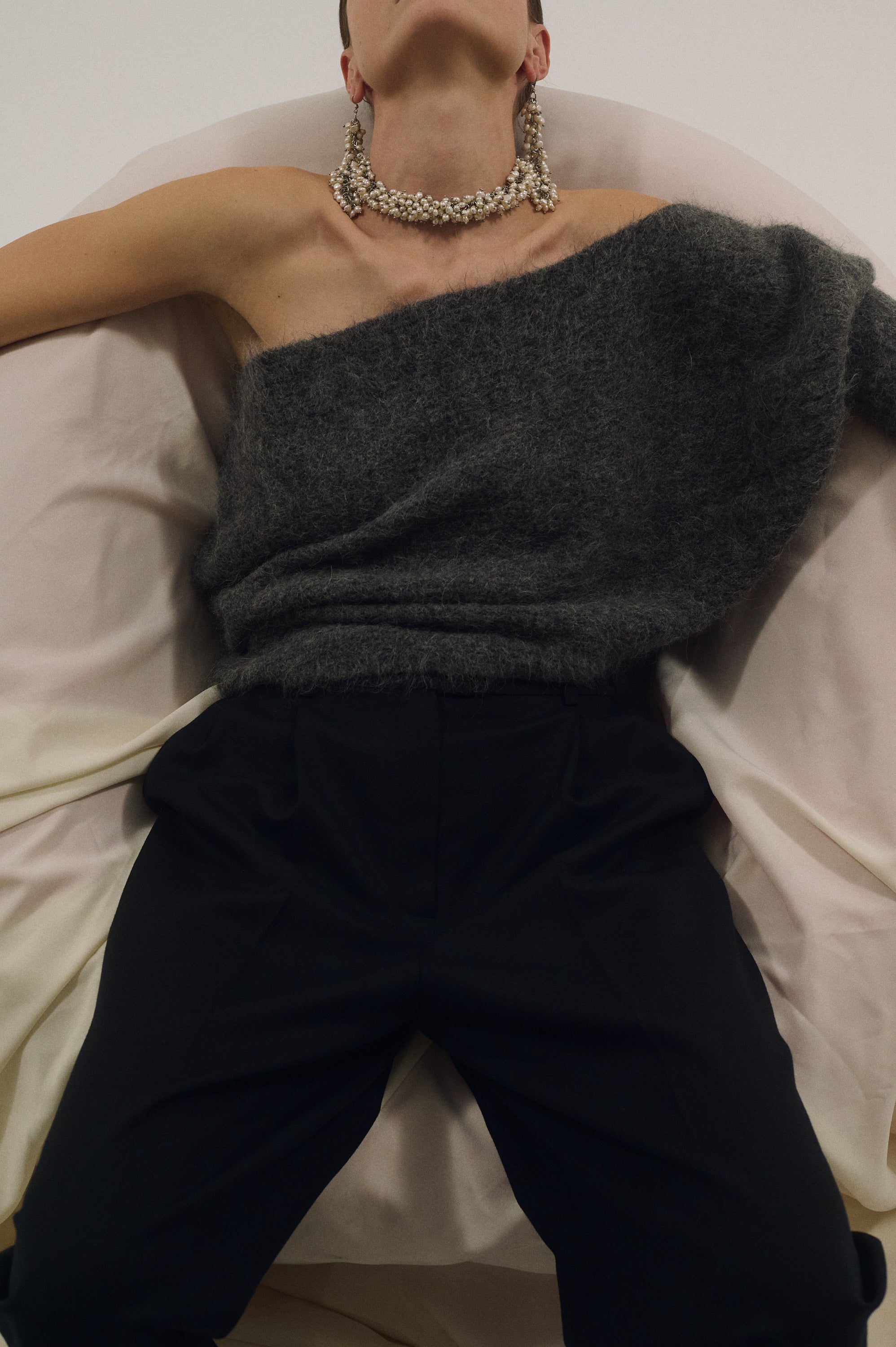 Person wearing massive pearl choker, dark gray sweater and black pants on a white couch.