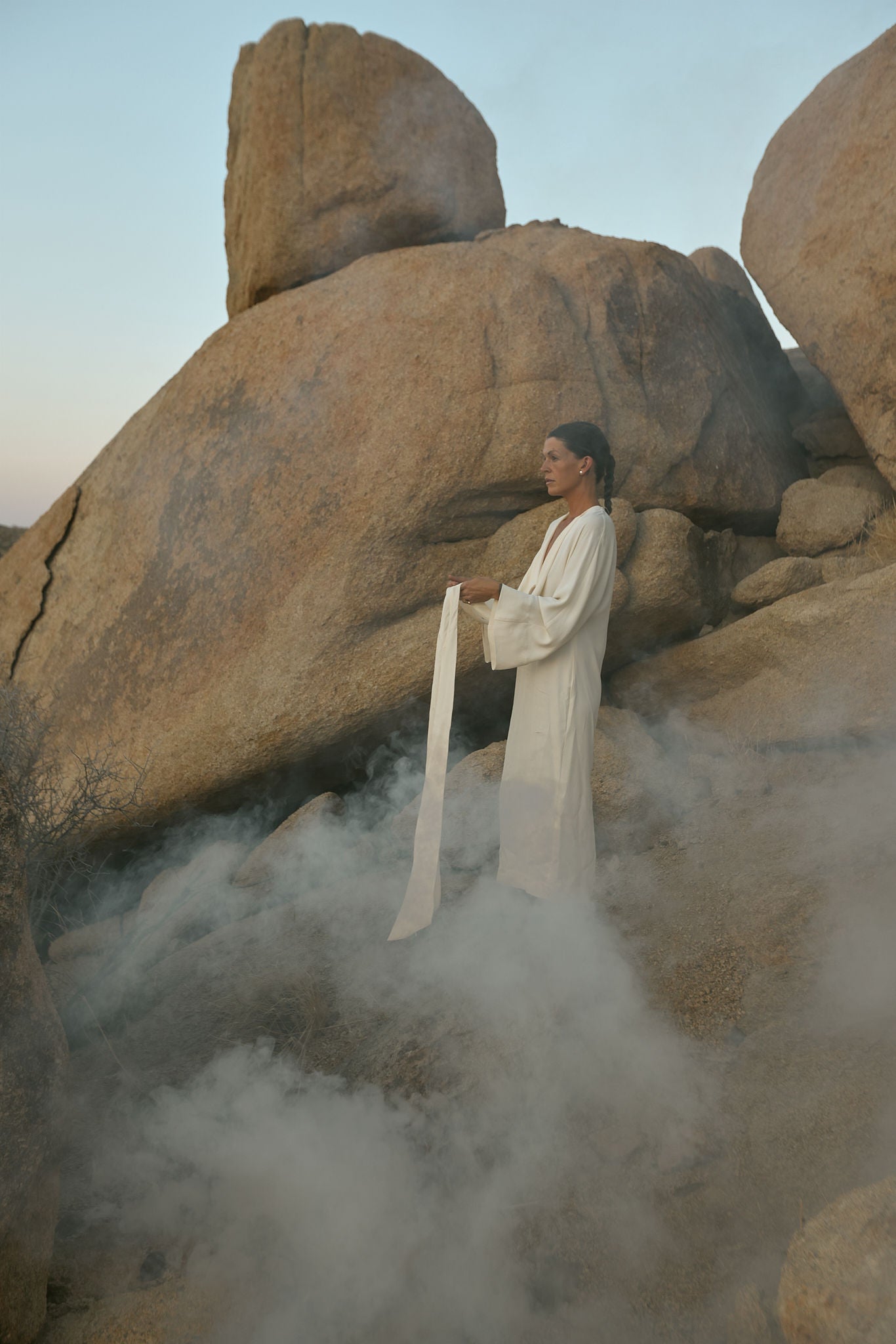 Person in a white robe standing among large rocks with mist with minimalist pearl jewelry