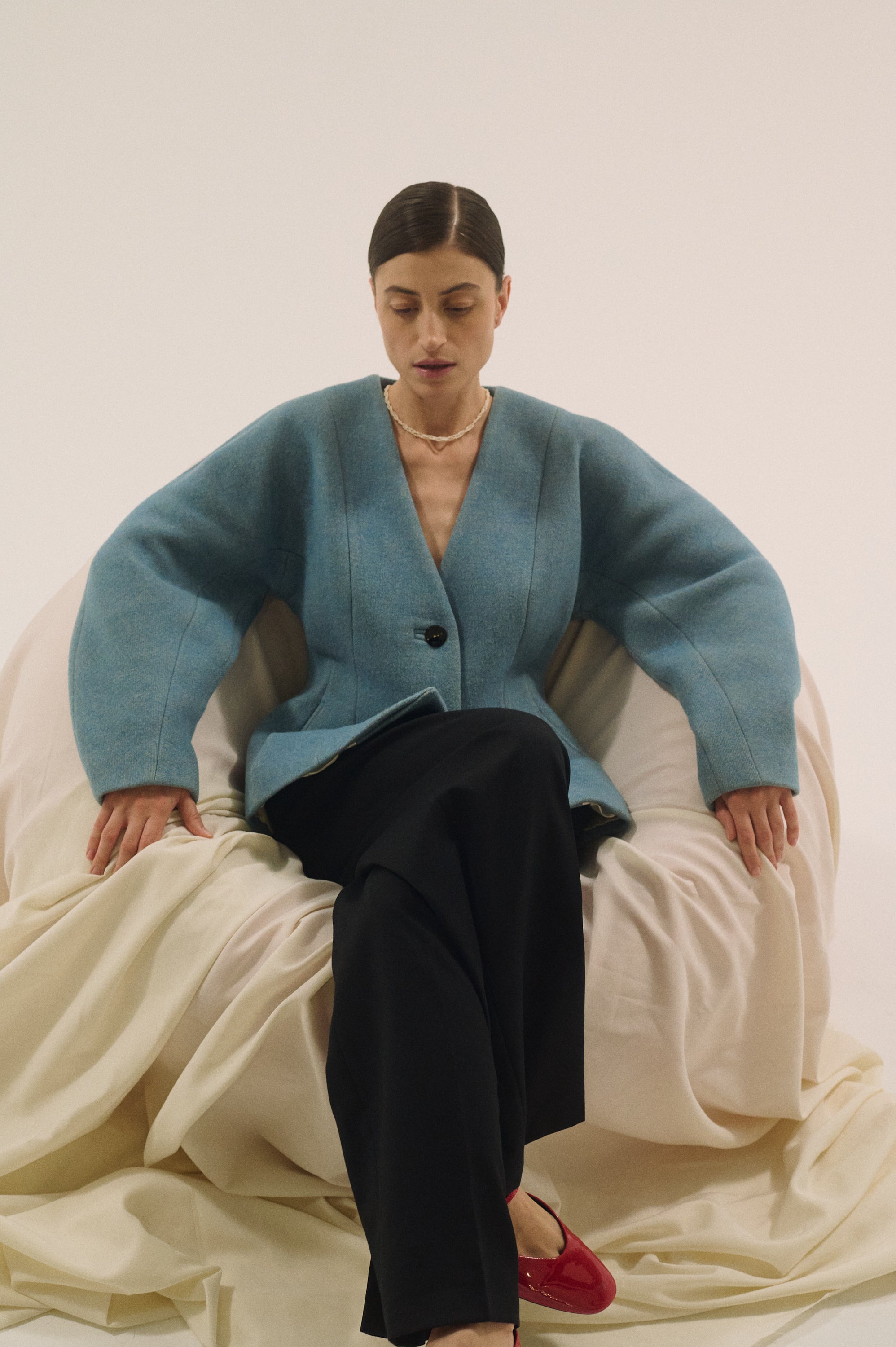 Full-body shot of a woman in a blue jacket and black trousers sitting on draped fabric, wearing a layered pearl choker.