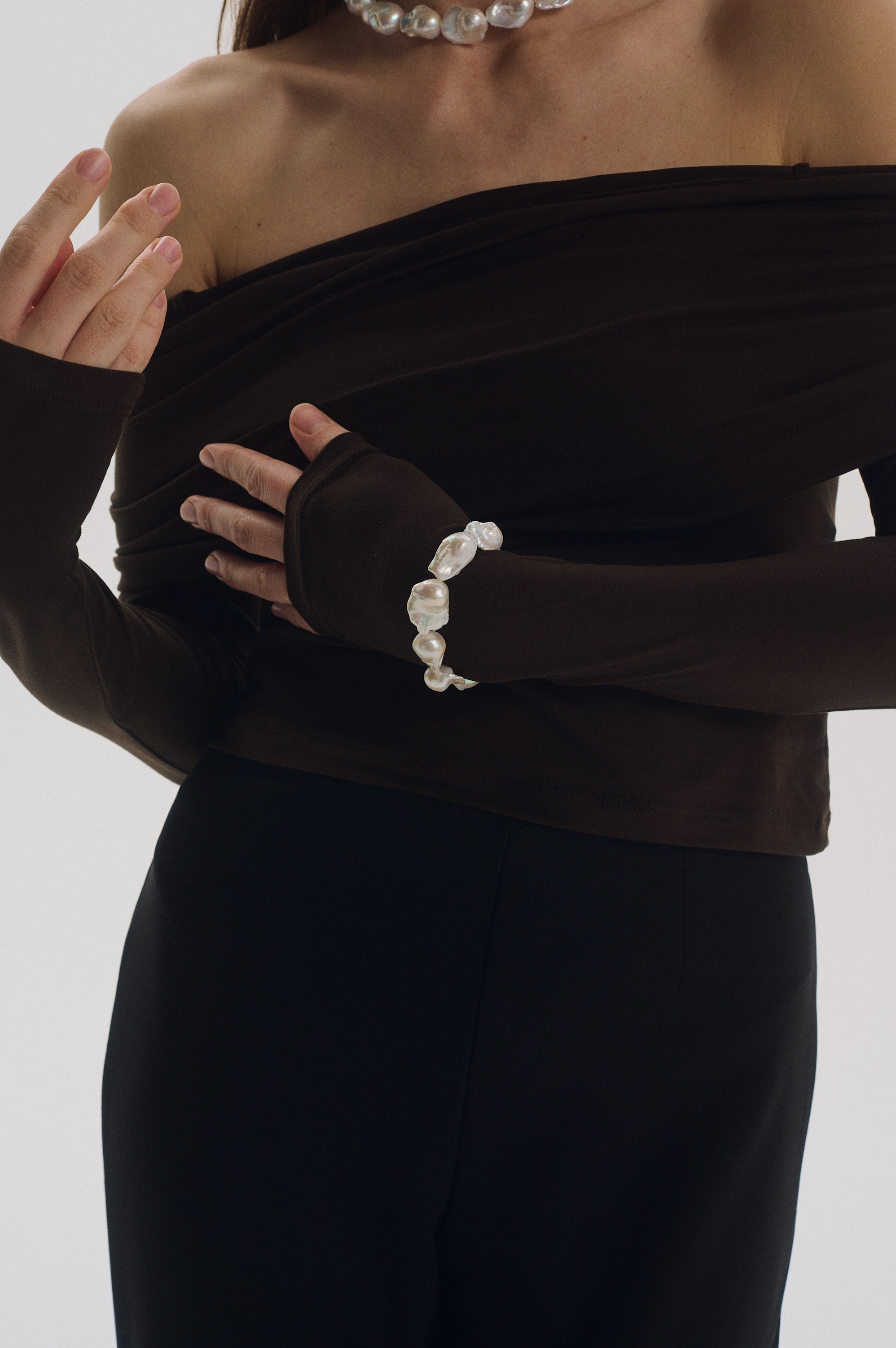 Person wearing a real pearl bracelet, dark brown off-shoulder top with matching pants on a light gray background