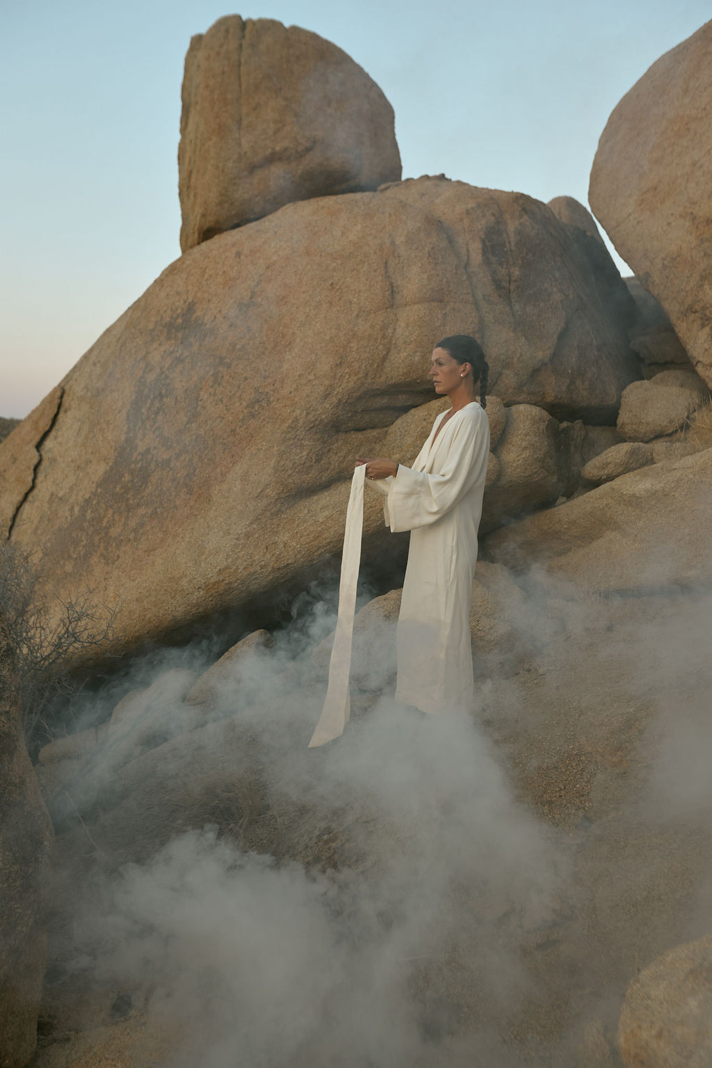 Person in a white robe standing among large rocks with mist with minimalist pearl jewelry