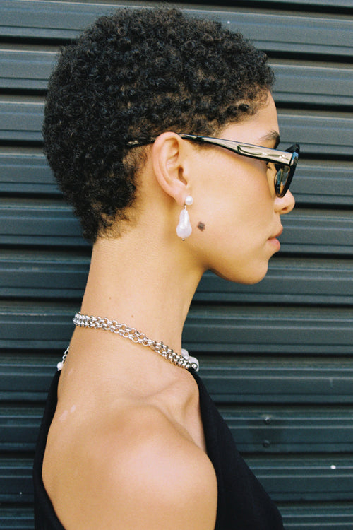 Side profile of a person with short natural hair wearing sunglasses and a necklace against a metallic background